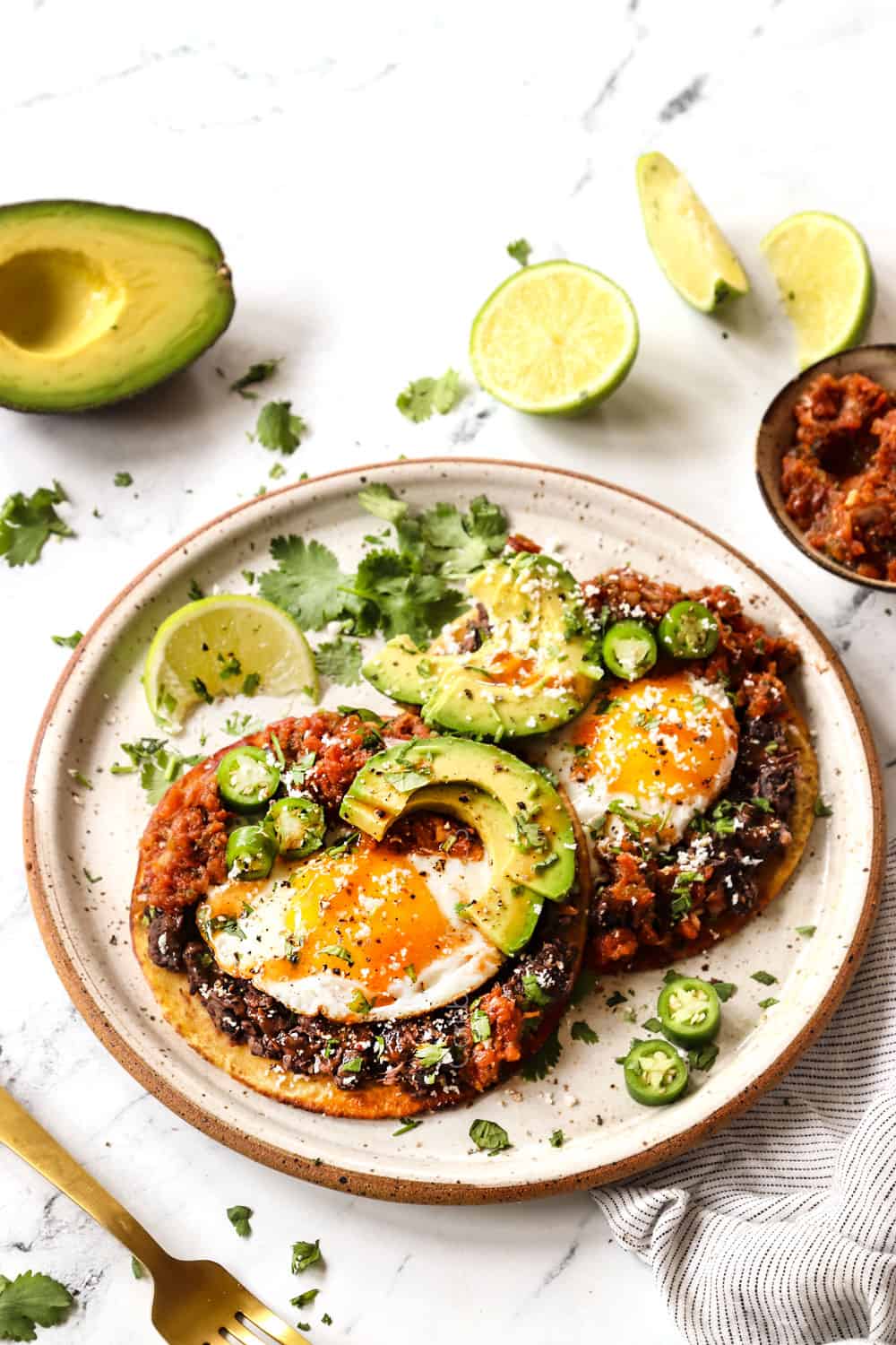 two huevos rancheros on a plate topped with avocados, sliced avocados, Cotija cheese and hot sauce