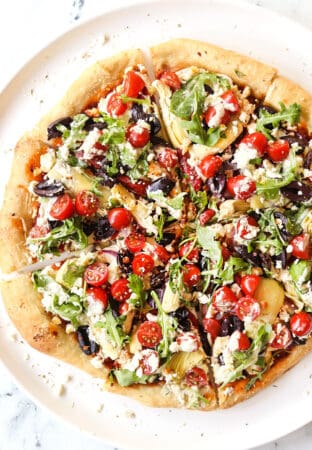 showing how to make Greek pizza recipe by adding arugula, tomatoes, cucumbers and tzatziki to the top of the baked pizza