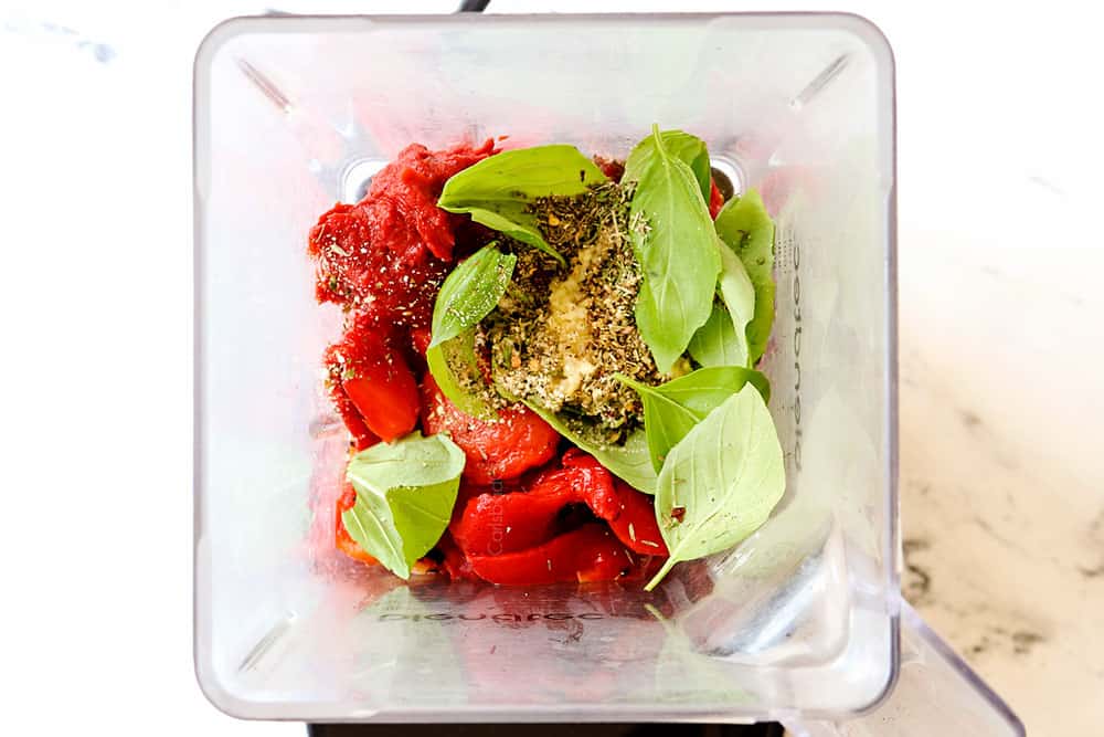 top view of showing how to make roasted red pepper sauce by adding roasted red peppers, garlic, basil, tomato paste, oregano, parsley, thyme and red pepper flakes to a blender