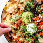 showing how to serve homemade loaded beef nacho recipe by topping chips with guacamole, sour cream, pico and olives with a hand taking a chip