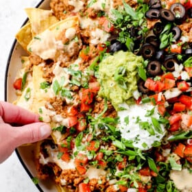 showing how to serve homemade loaded beef nacho recipe by topping chips with guacamole, sour cream, pico and olives with a hand taking a chip