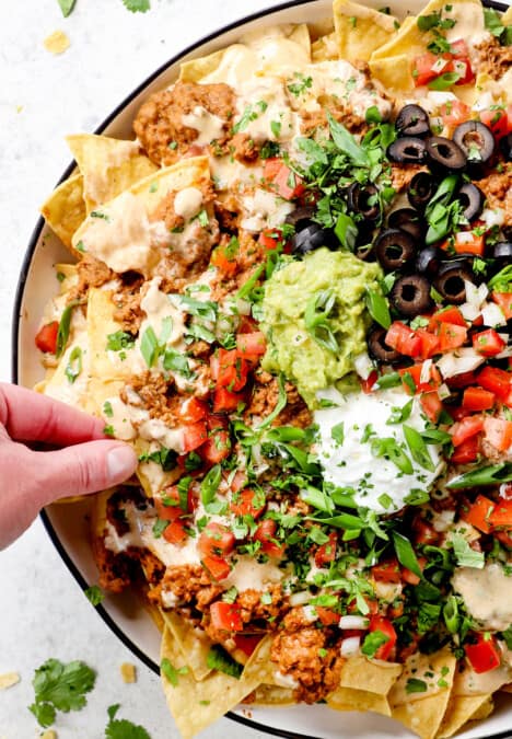 showing how to serve homemade loaded beef nacho recipe by topping chips with guacamole, sour cream, pico and olives with a hand taking a chip