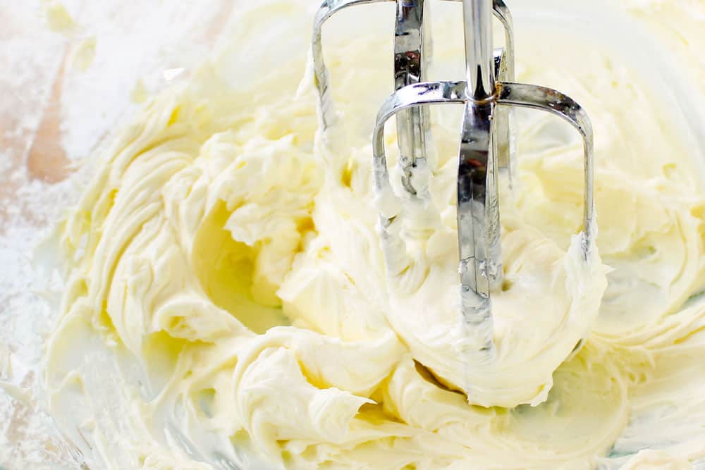showing how to make Ooey Gooey Butter Cake recipe by beating cream cheese until smooth and lump free in a glass mixing bowl with hand held mixer