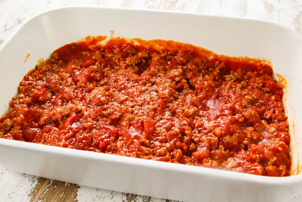 showing how to make stuffed shells recipe by adding marinara with meat to the bottom of a 9x13 pan