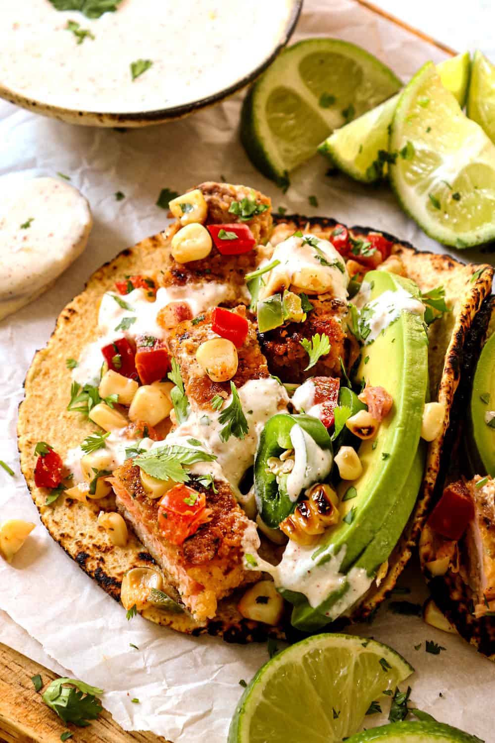 showing how to make fish tacos by layering salmon, corn salsa, avocados and taco sauce on corn tortilla