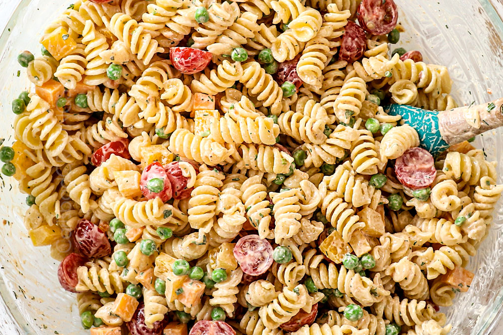 showing how to make bacon ranch pasta salad by tossing the pasta salad with half of the dressing before refrigerating