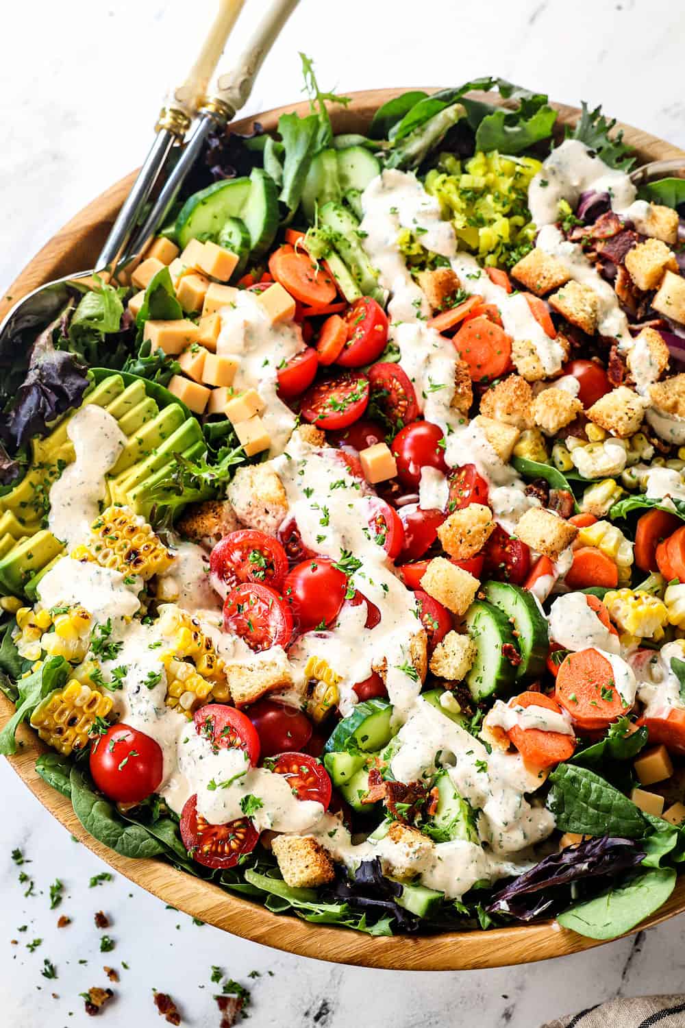 showing ingredients for garden salad with lettuce, tomatoes, cucumbers, carrots, avocados, pepperoncini, red onion, croutons, cheddar cheese and bacon drizzled with ranch