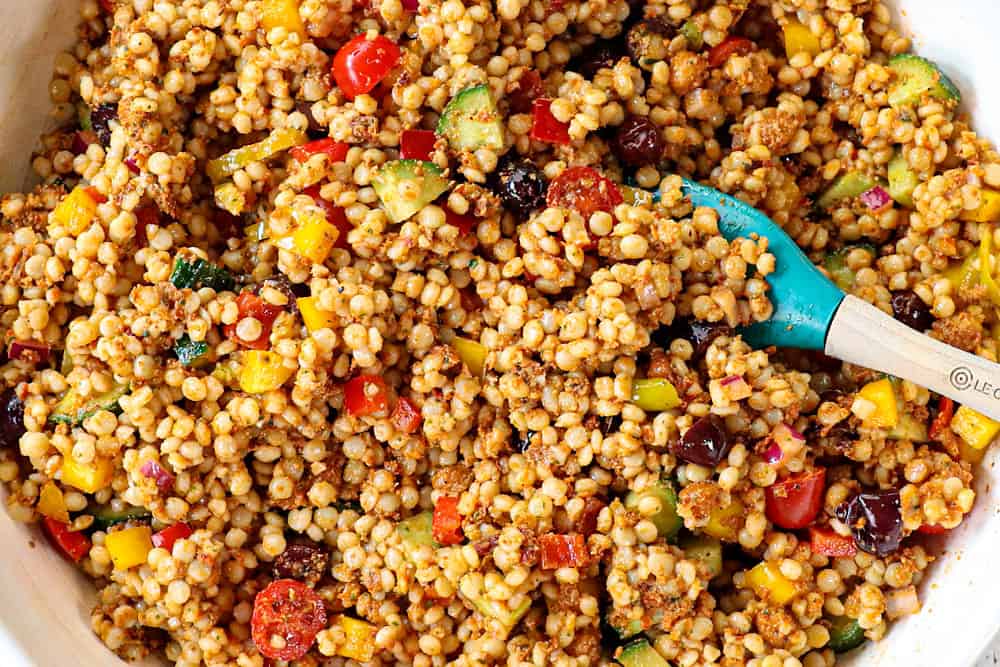 showing how to make couscous salad by tossing the pearl couscous with vegetables and dressing in a white bowl