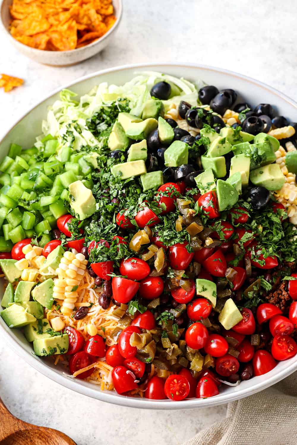 side view showing how to make taco pasta salad recipe by adding pasta, ground beef, black beans, tomatoes, avocados, olives, lettuce and cheese to a white bowl