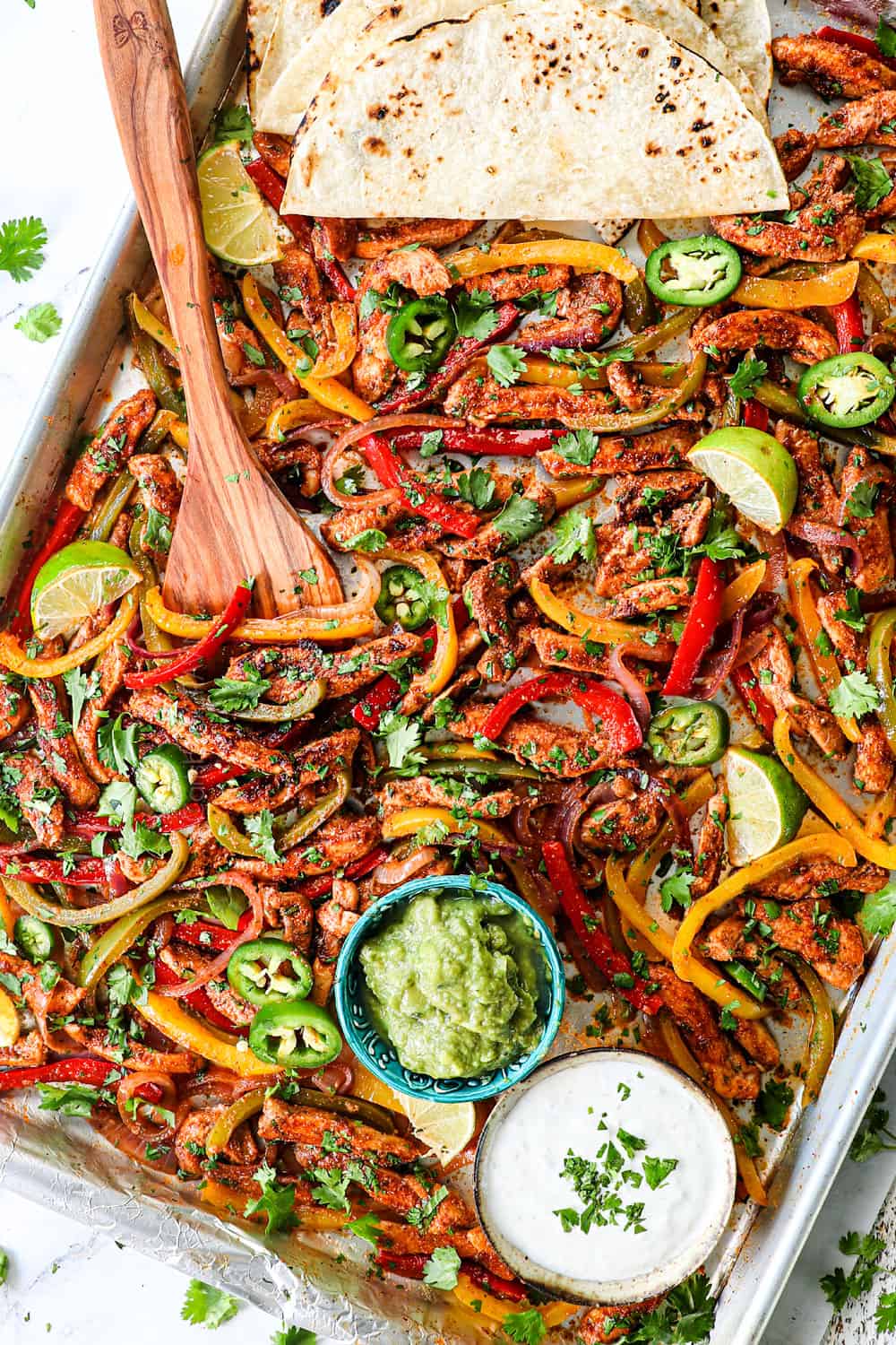 showing how to serve sheet pan chicken fajitas by adding tortillas, guacamole and sour cream to the serving platter