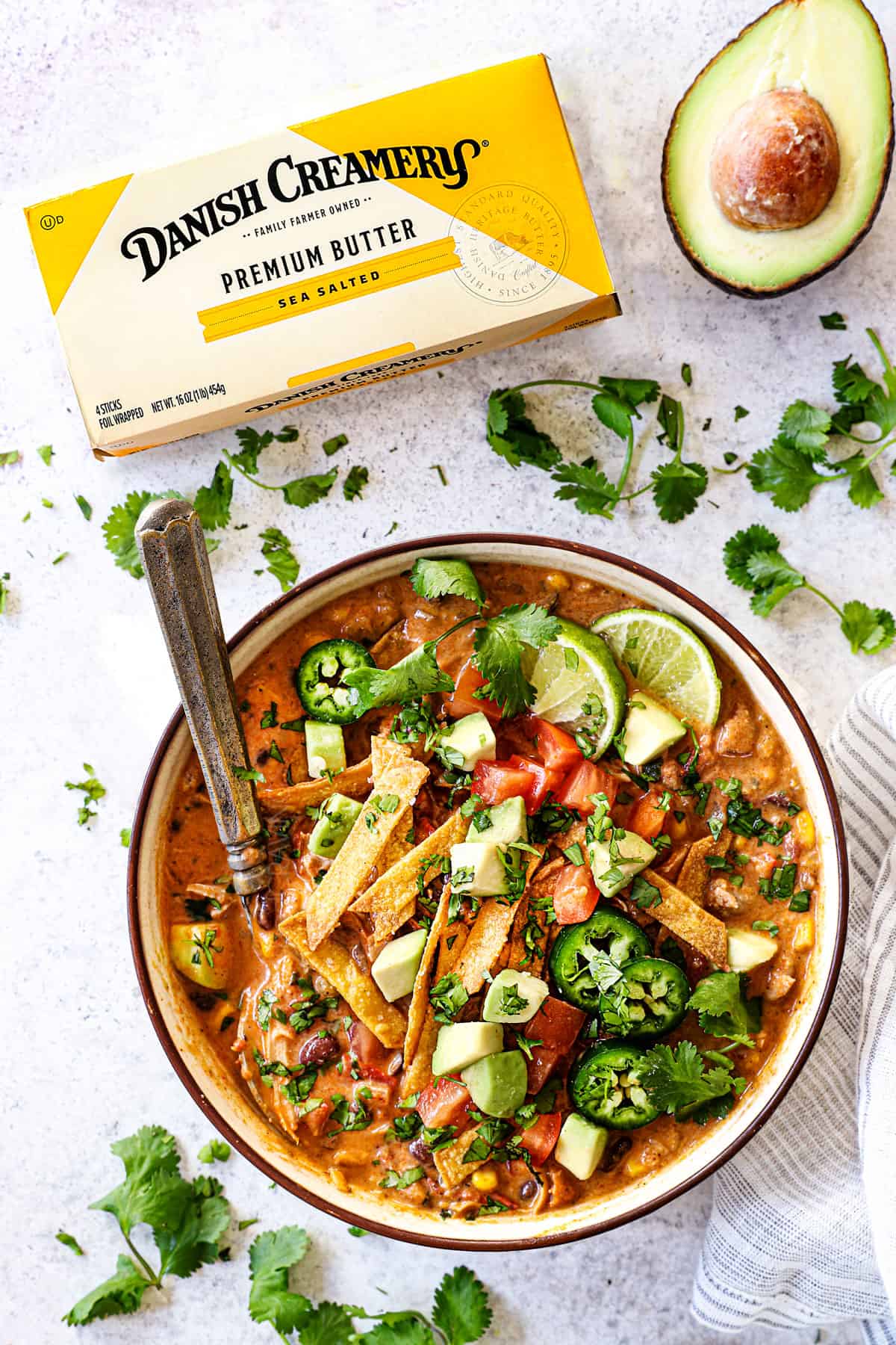 top view showing how to serve chicken tortilla soup by adding to a bowl and topping with tortilla strips, avocado, cilantro, jalapeno, fresh limes and tomatoes