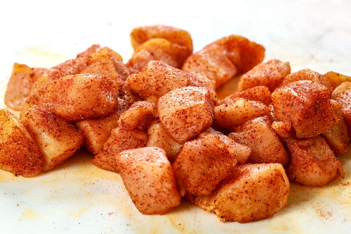 showing how to make chicken bites recipe by tossing cubed chicken breasts with seasonings on a white cutting board