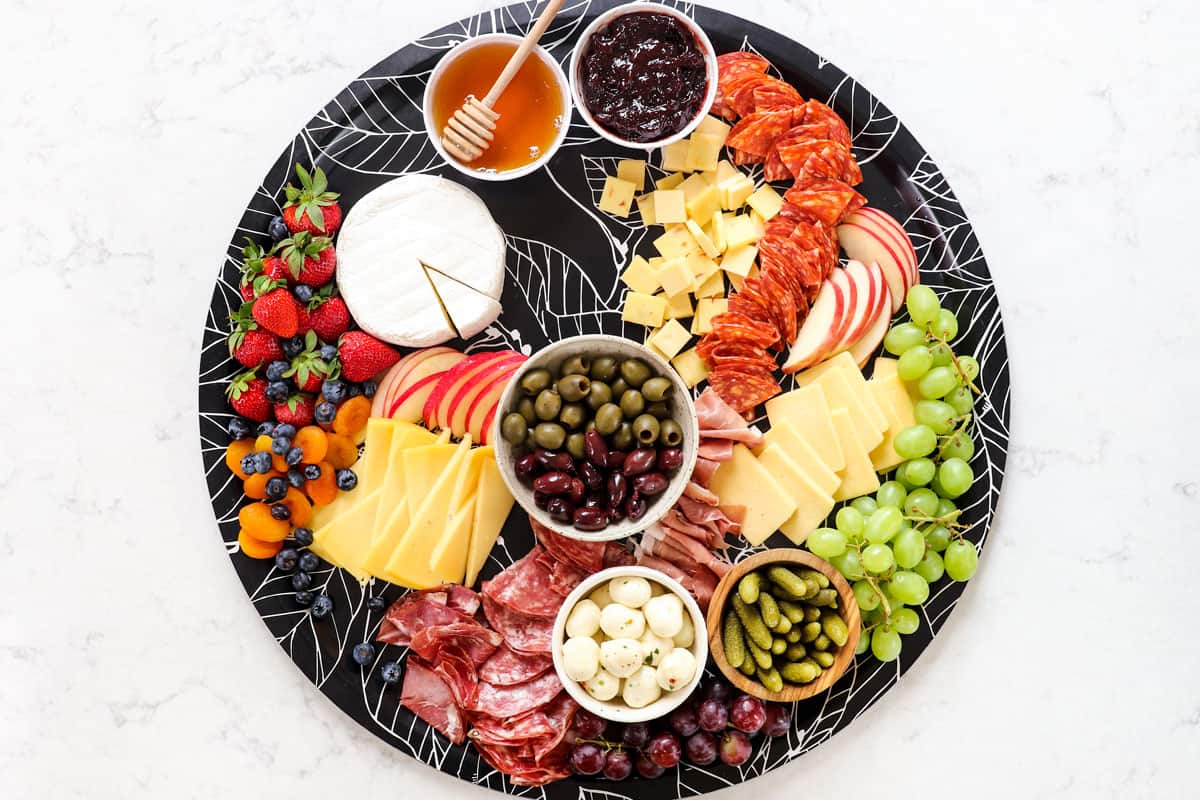 showing how to make a charcuterie board by adding the fruit (strawberries, grapes, blueberries, dried apricots) to the board by the cheese