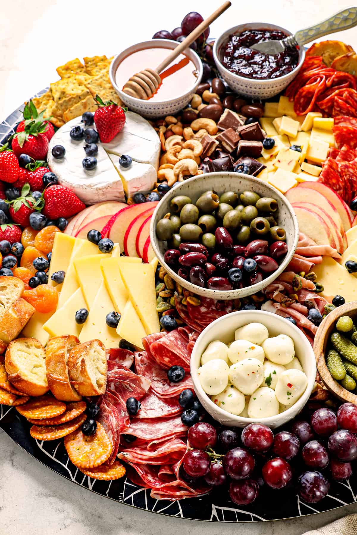 a meat and cheese charcuterie board with salami, prosciutto, Havarti, Gouda, brie, goat cheese, grapes, strawberries, blueberries, apples, honey, jam and nuts to a board