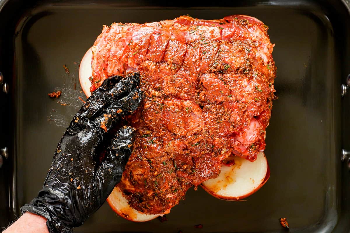 showing how to cook leg of lamb by rubbing the top side of the lamb roast with a garlic, rosemary wet rub and placing it in a roasting pan with onions and garlic