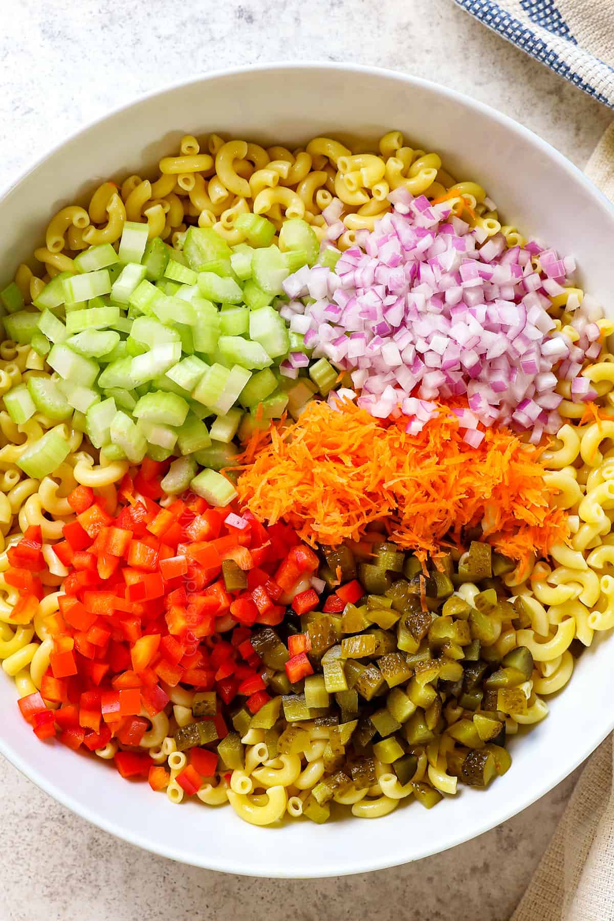 top view showing ingredients of macaroni salad recipe in a bowl: macaroni, celery, red onions, pickles, shredded carrots