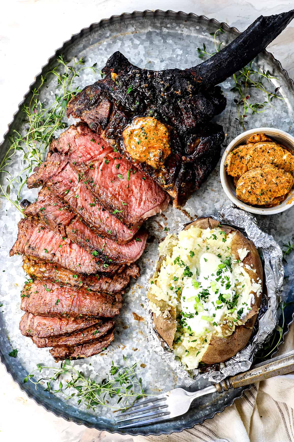 showing how to serve tomahawk steaks on a serving platter with baked potatoes, compound butter and fresh herbs