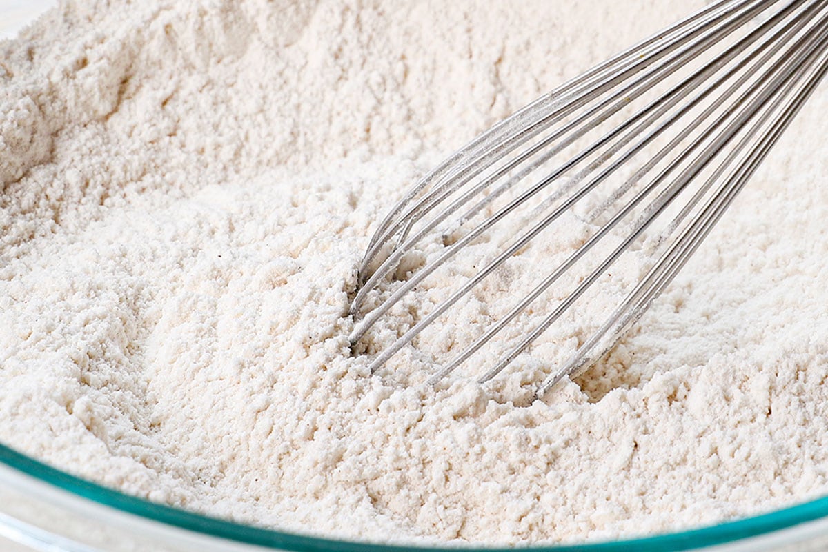 showing how to make fluffy pancakes recipe by whisking the flour, sugar, baking powder, cinnamon, and salt in a large mixing bowl.