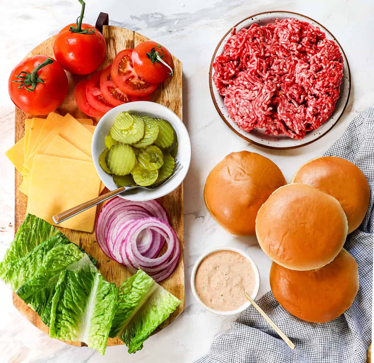 top view showing cheeseburger ingredients: ground beef, hamburger buns, cheese slices, tomatoes, pickles, red onion, lettuce, burger sauce