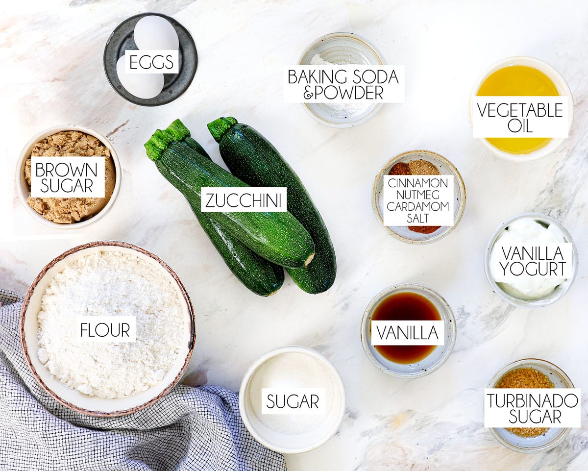 top view of zucchini bread ingredients: shredded zucchini, flour, brown sugar, granulated sugar, vanilla extract, eggs, baking powder, baking soda, vanilla yogurt, vegetable oil
