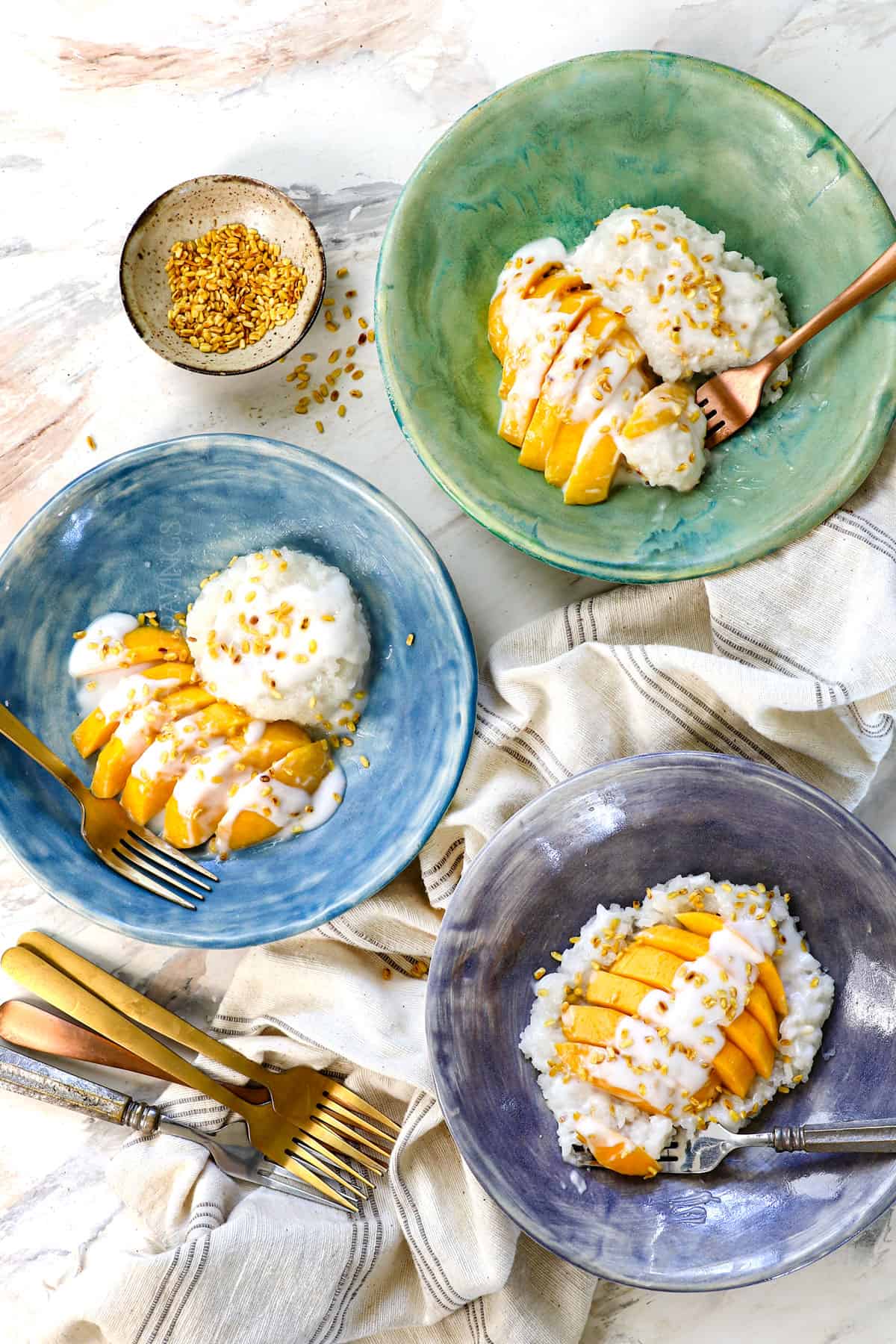 top view of mango sticky rice recipe served in three bowls in different variations: one with the rice in a pile with mango on the side, one with the mango on top of the rice, and one with the mango to the side of rice in a long shape