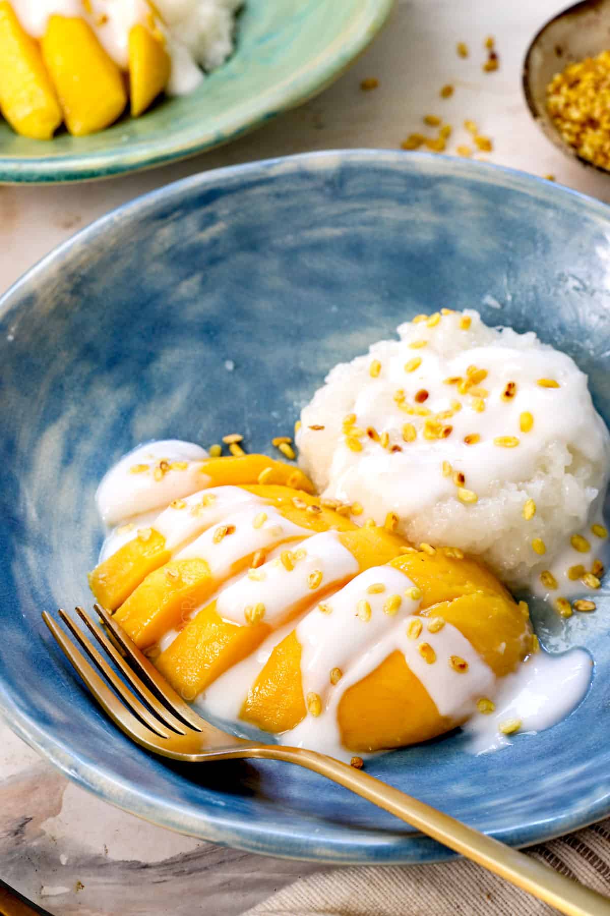 side view of sticky mango rice being served in a blue shallow dish with a gold fork showing how creamy the coconut sauce is
