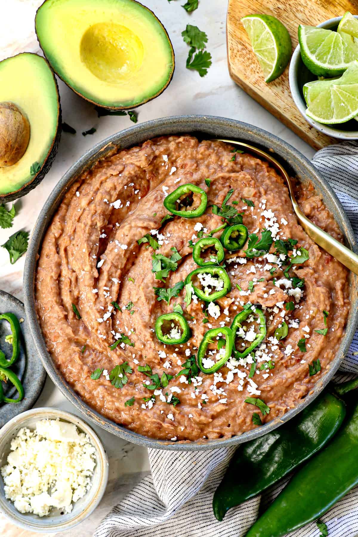 top view of refried bean recipe being with avocados, cilantro, limes, jalapenos, and cotija cheese on the side for serving