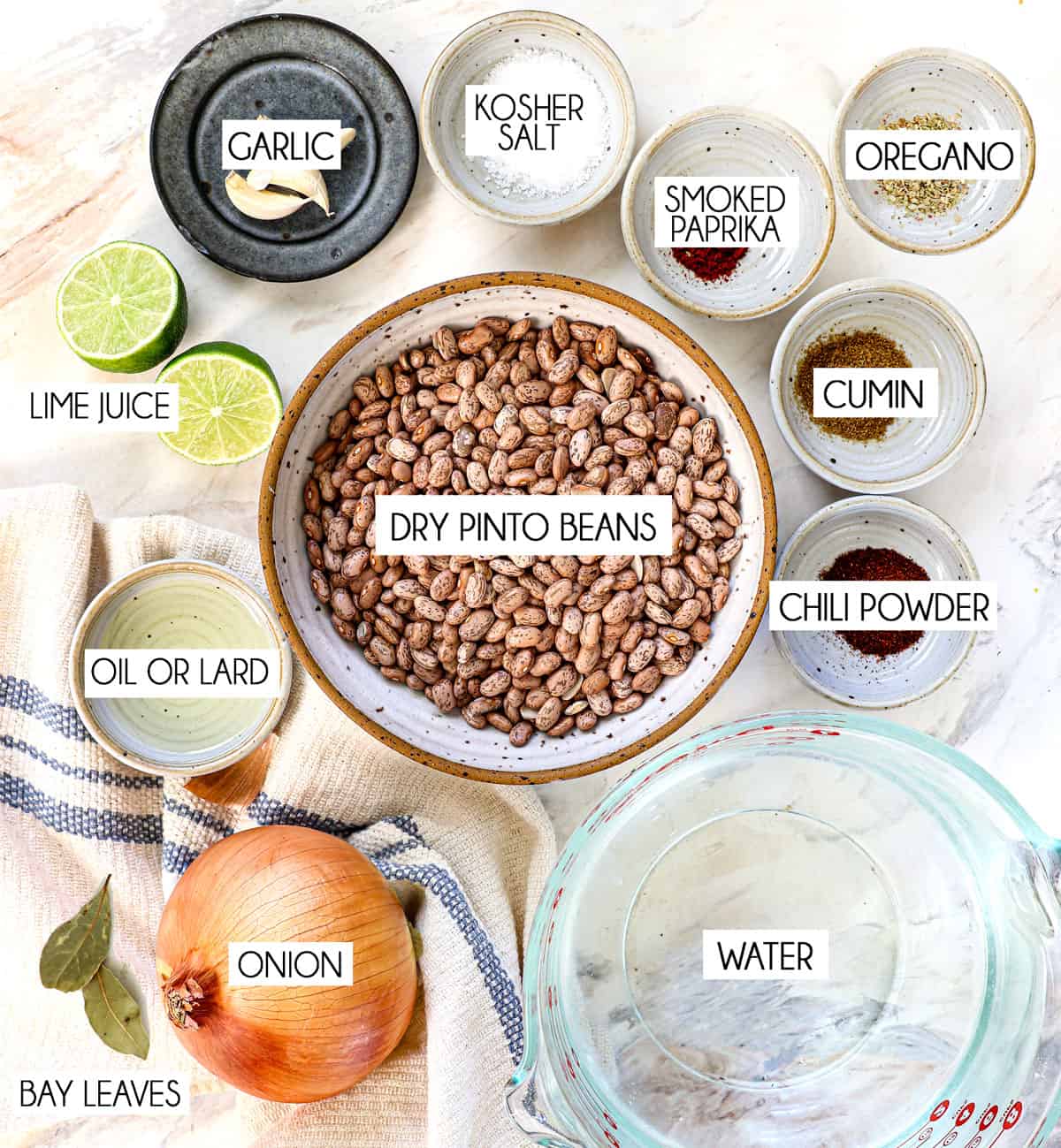 top view of refried beans recipe ingredients: dry pinto beans, onion, garlic, water, ground cumin, chili powder, dried oregano, smoked paprika, kosher salt, lime juice and vegetable oil or lard