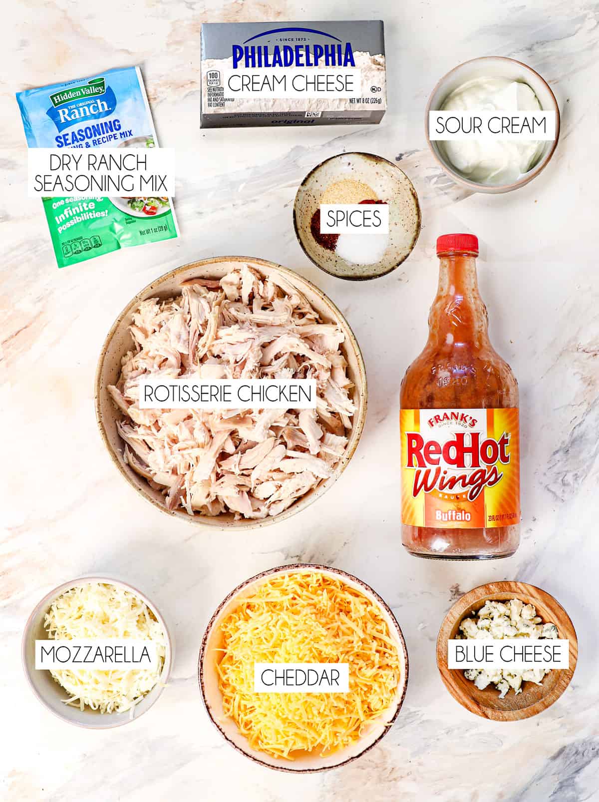 top view of buffalo chicken dip ingredients: Frank's Buffalo Wings sauce, cream cheese, shredded chicken, ranch seasoning mix, sour cream cheddar cheese, mozzarella cheese, blue cheese, and spices