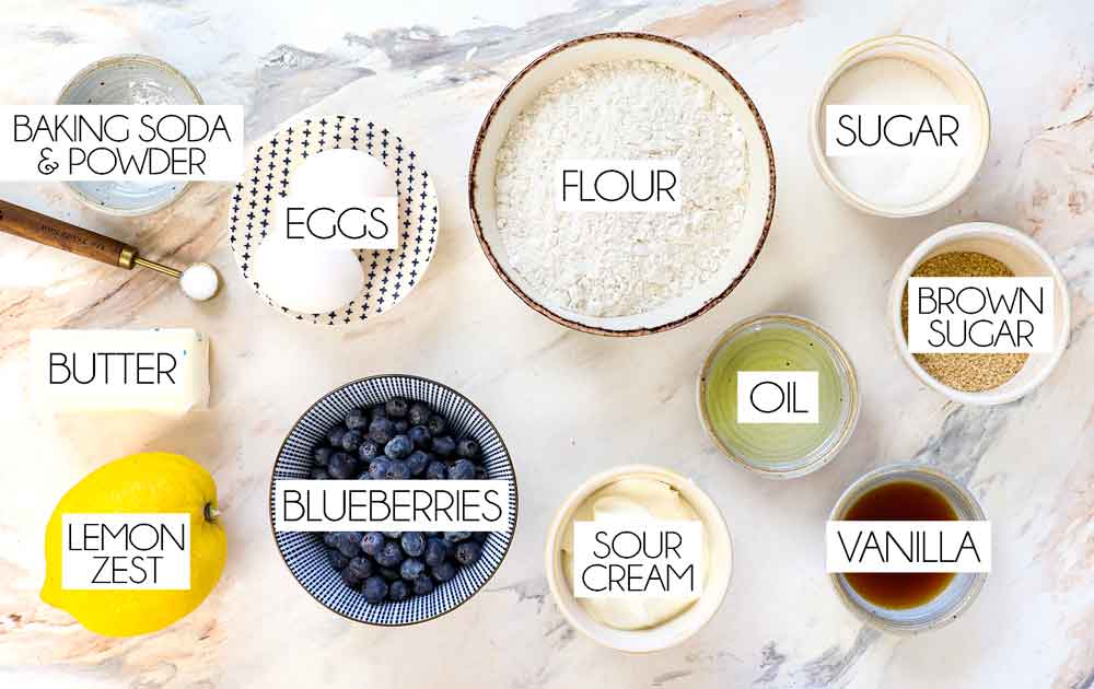 top view of blueberry muffin recipe ingredients: flour, blueberries, sugar, eggs, baking soda, baking powder, lemon zest, vegetable oil, butter, sour cream, vanilla