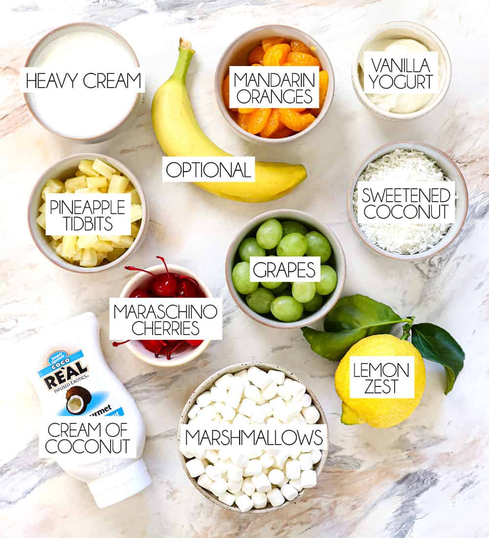 top view of ambrosia salad recipe ingredients: maraschino cherries, pineapple tidbits, mandarin oranges, grapes, marshmallows, shredded coconut, heavy cream, vanilla yogurt, lemon zest, cream of coconut