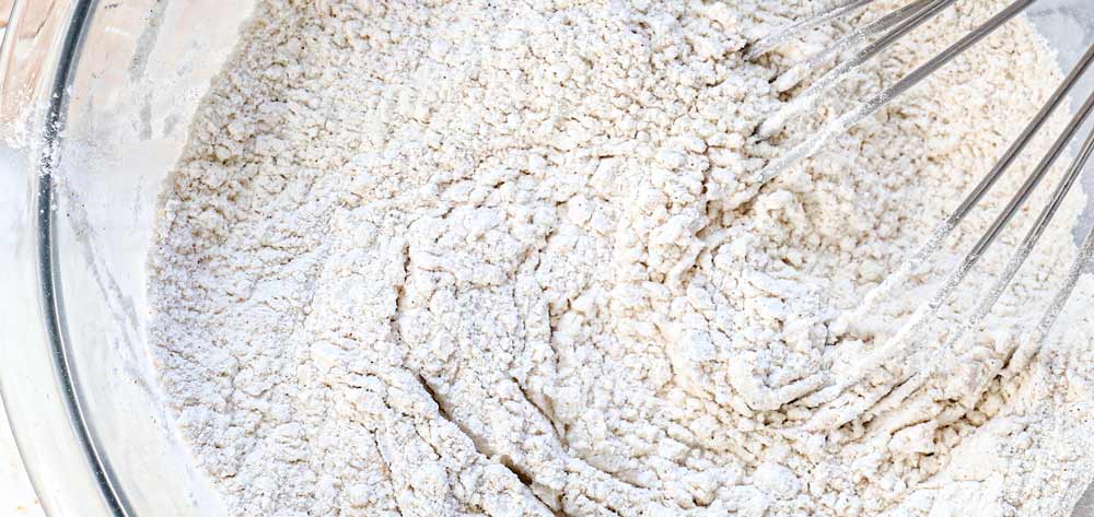 showing how to make blueberry muffins by whisking the flour, baking powder, baking soda, and ground cinnamon together in a bowl