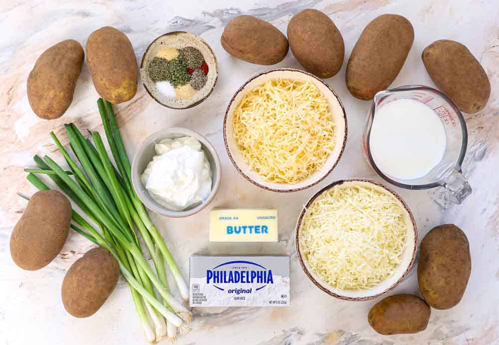 top view of mashed potato casserole ingredients: Russet potatoes, milk, sour cream, cheeses, spices, green onions, butter, cream cheese